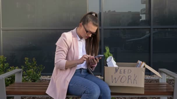 Нужна работа. На скамейке возле офиса сидит очень расстроенная молодая женщина. В ее руках коробка с личными вещами и сотовым телефоном. Женщина пытается найти работу через Интернет . — стоковое видео