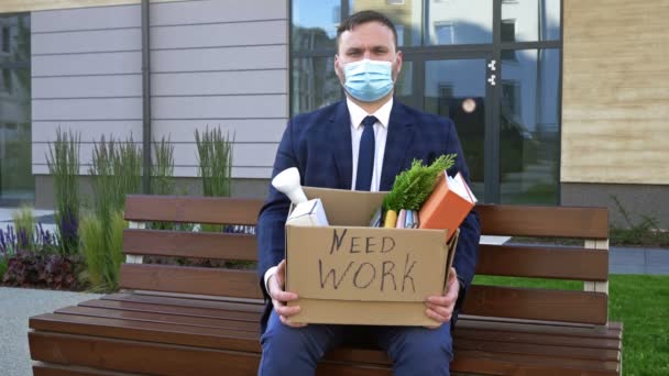 Рядом с офисом сидит очень расстроенный мужчина средних лет в медицинской маске. В его руках коробка с личными вещами. На коробке написано "Нужна работа". Безработица в результате пандемии COVID-19 . — стоковое видео