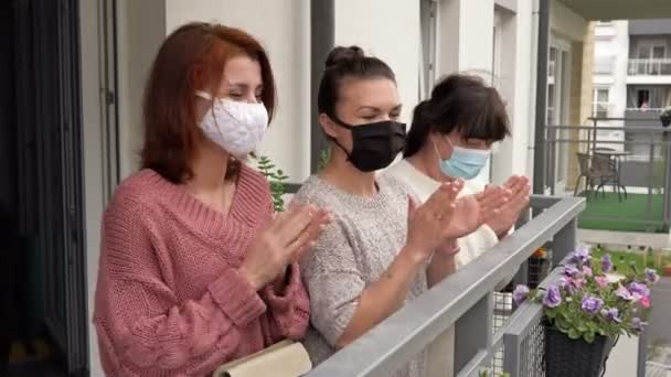 Drei Frauen in medizinischen Masken, die auf dem Balkon eines Mehrfamilienhauses stehen, winken fröhlich mit den Händen. — Stockvideo