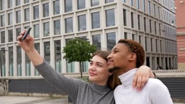 Genç Afrikalı bir adam ve beyaz bir kız şehir meydanında durup selfie çekiyorlar. Farklı ten renkleri olan insanlar arasındaki ilişkiler.