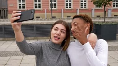 Genç Afrikalı bir adam ve beyaz bir kız şehir meydanında durup selfie çekiyorlar. Farklı ten renkleri olan insanlar arasındaki ilişkiler.