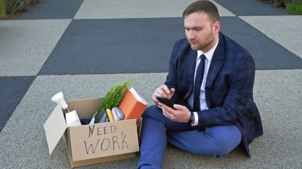 Triste manager d'âge moyen assis sur le sol avec ses effets personnels après avoir perdu son emploi. L'homme essaie de trouver du travail en utilisant Internet. — Video