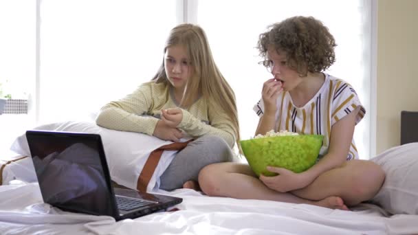 Two teenage girlfriends watching a horror movie at home sitting on the bed and eating popcorn. They are both scared and fun. — Stock Video