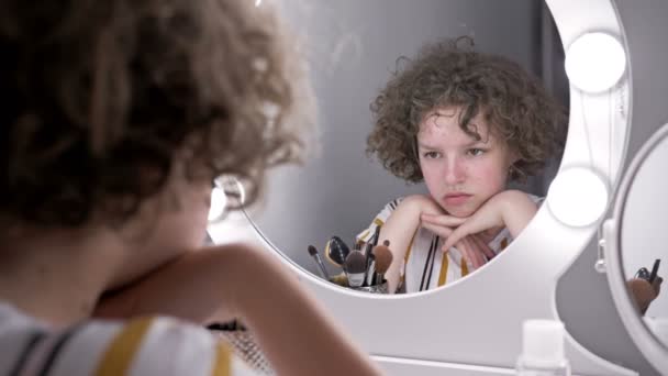 Το κορίτσι κοιτάζει προσεκτικά στον καθρέφτη το πρόσωπό της. Είναι πολύ αναστατωμένη από την εφηβική ακμή.. — Αρχείο Βίντεο