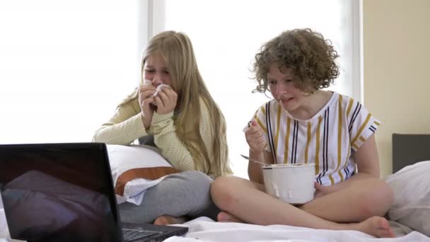 自宅で劇的な映画を見て、ベッドの上に座ってアイスクリームを食べる十代の年齢の2人のガールフレンド。かれらは感動して泣き，. — ストック動画