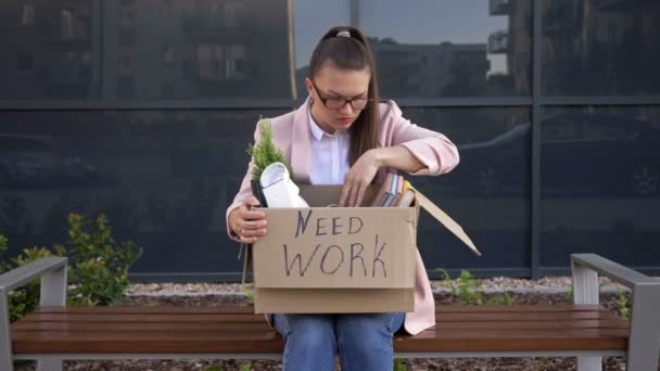 Jovem que é deixado sem trabalho está segurando um cartaz precisa de trabalho. Ela está sentada em um banco sob o escritório em protesto . — Vídeo de Stock