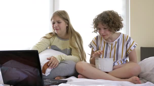 Dos amigas adolescentes viendo películas dramáticas en casa, sentadas en la cama y comiendo helado. Ambos se mueven y lloran. — Vídeo de stock