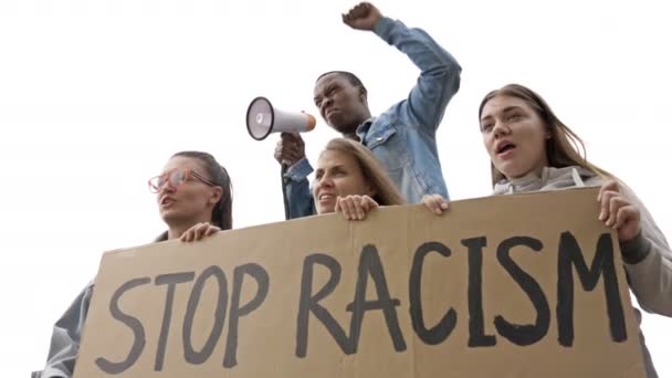 Grupa młodych ludzi posiada plakat STOP RACISM. Młody protestujący o ciemnej skórze wykrzykuje slogany do megafonu. — Wideo stockowe