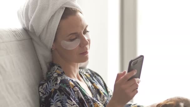 Middelbare vrouw na een douche zit op een bank en kijkt naar het scherm van een mobiele telefoon. Haar huisdier heeft de aandacht van de gastvrouw nodig.. — Stockvideo