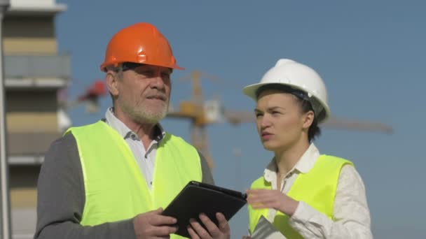 Idoso com um tablet nas mãos e uma jovem mulher em um canteiro de obras. Construtores se divertir discutindo questões de trabalho . — Vídeo de Stock