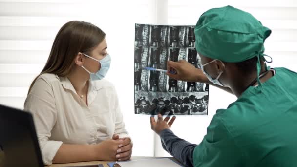 Černý doktor vysvětluje pacientovi rentgen a dělá doporučení. Doktor a pacient v lékařských maskách. Covid-19. — Stock video