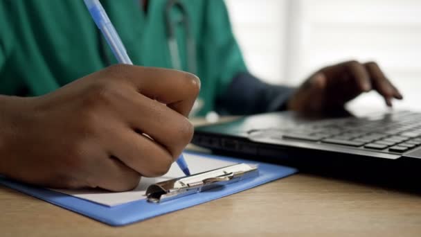 Médecin noir travaille à un ordinateur et prend des notes dans son bureau. Gros plan des mains du médecin. — Video