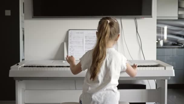 5-6-летняя девочка садится перед белым цифровым пианино и нажимает клавиши. Вид сзади . — стоковое видео