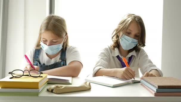 Uczennica i uczennica siedzą przy tym samym biurku podczas lekcji. Na twarzach nastolatków, maski ochronne. Zapobieganie epidemii covid-19. — Wideo stockowe