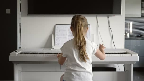 5-6 yaşlarında bir kız beyaz dijital bir piyanonun önünde oturuyor ve tuşlara basıyor. Arka plan. — Stok video