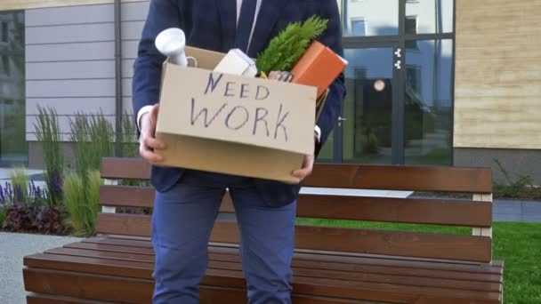 Hospodářská krize. Velmi rozrušený muž středního věku sedí na lavičce poblíž kanceláře. V jeho rukou je krabice s osobními věcmi. Na krabici je napsáno NEED WORK. — Stock video