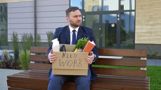 経済危機。オフィスの近くのベンチには非常に動揺した中年の男が座っている。彼の手には個人的な持ち物の箱がある。箱の上には仕事が必要だと書いてある. — ストック動画