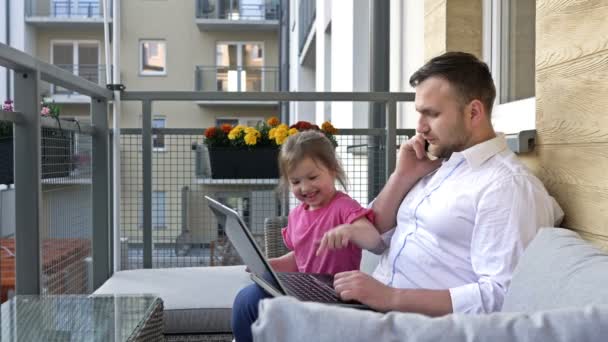 Travaillez à distance. Le jeune homme travaille sur le balcon à la maison. La petite fille veut jouer avec son père et l'empêche de travailler. — Video