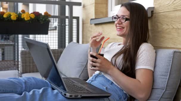 Mujer joven está feliz de comunicarse con alguien en línea. Ella está cómodamente sentada en el balcón de su apartamento . — Vídeos de Stock
