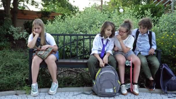 Трое школьников сидят на скамейке во дворе школы. Дети смеются над девушкой, сидящей рядом с ним. Школьные издевательства. — стоковое видео