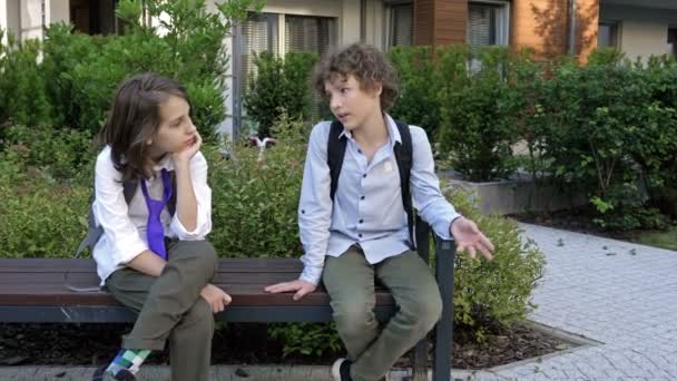 İki okul çocuğu okul bahçesinde bir bankta oturuyorlar. Çocuklar hareketli bir şekilde konuşuyorlar. Okul arkadaşlığı. — Stok video