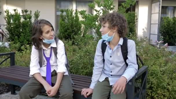 Två skolbarn sitter på en bänk och pratar livligt. Pojkarna tog av sig sina tråkiga medicinska masker.. — Stockvideo