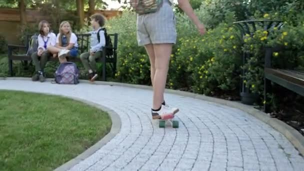 En tjej på en skateboard kör fram till klasskamrater som sitter på en bänk på skolgården. — Stockvideo