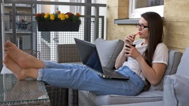 Attraktiv ung kvinde sidder på en balkon med en bærbar computer og en sodavand. Hun har det godt. Fordelene ved fjernarbejde. – Stock-video