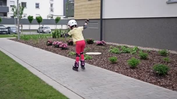 Маленька дівчинка катається на роликах у дворі багатоквартирного будинку. Дитина тільки починає освоювати роликові ковзани, тому часто падає . — стокове відео