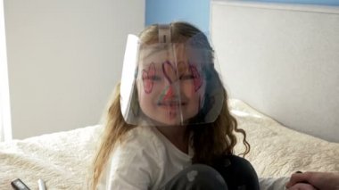 Koruyucu pleksiglas maskeli neşeli küçük kız. Maskenin üzerinde komik çizimler var. Birisi çocukla oynayan bir yetişkin..
