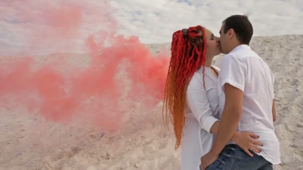 Pareja de amantes besándose sobre un fondo de arena blanca, nubes blancas y humo rojo. Fecha romántica . — Vídeo de stock