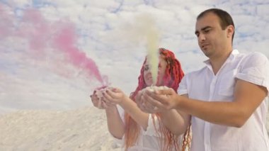 Güzel aşıklar beyaz kumların üzerinde duruyor. Ellerinde renkli duman vardı. Romantik tarih beyaz bulutların arkaplanına karşı.