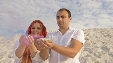 Güzel aşıklar beyaz kumların üzerinde duruyor. Ellerinde renkli duman vardı. Romantik tarih beyaz bulutların arkaplanına karşı.