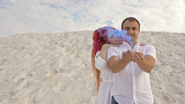 Hermosa chica abraza y besa a su amante. El hombre tiene humo azul. Fecha romántica aislada contra el telón de fondo de arena blanca y un cielo con nubes blancas . — Vídeos de Stock