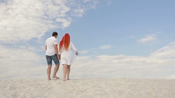 Um homem e uma mulher caminham na areia branca de mãos dadas. Contra o fundo do céu azul e nuvens brancas. Visão traseira . — Vídeo de Stock