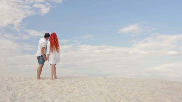L'uomo e la donna stanno sulla spiaggia sabbiosa tenendosi per mano e ammirando le nuvole. — Video Stock