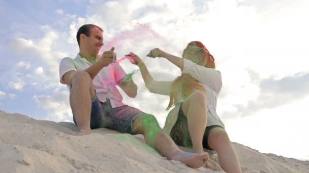 Jóvenes y hermosos amantes sentados sobre arena blanca se rocían unos a otros con coloridos polvos Holi. Las tradiciones indias en Europa. — Vídeo de stock