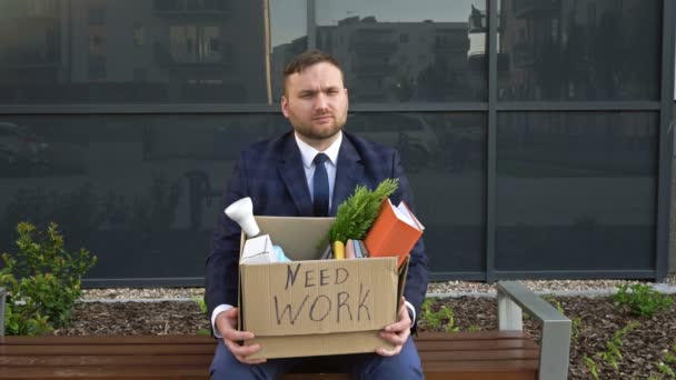 Crise económica. No banco perto do escritório fica um homem de meia-idade muito chateado. Em suas mãos está uma caixa com pertences pessoais. Na caixa diz NECESSÁRIO TRABALHO . — Vídeo de Stock