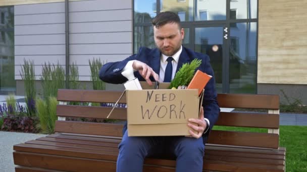 No banco perto do escritório fica um homem de meia-idade muito chateado. Em suas mãos está uma caixa com pertences pessoais. Na caixa diz NECESSÁRIO TRABALHO . — Vídeo de Stock