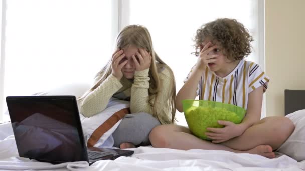 Two teenage girlfriends watching a horror movie at home sitting on the bed and eating popcorn. They are both scared and fun. — Stock Video