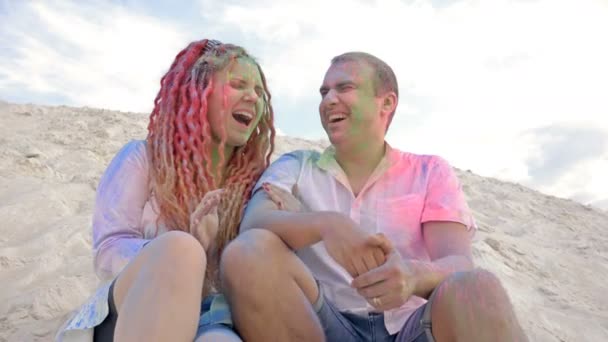 Retrato de um jovem casal alegre sentado na areia branca. As caras e a roupa pintam-se em cores brilhantes. Festival Holi . — Vídeo de Stock