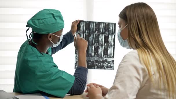 El médico negro explica al paciente una radiografía y hace una recomendación. Médico y paciente con máscaras médicas. Covid-19. — Vídeo de stock