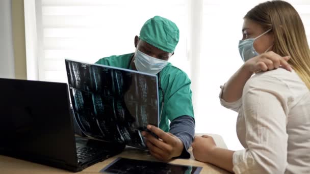 Kaukasische Patientin berät sich mit einem jungen afrikanischen Arzt über Rückenschmerzen. Arzt und Patient in medizinischen Masken. Covid-19. — Stockvideo