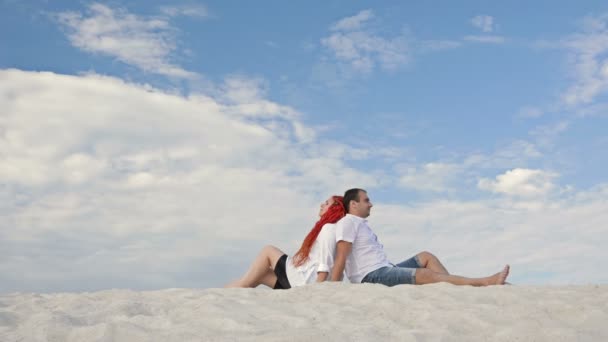 A boldog szerelmespár háttal ül a fehér homokon a kék ég felé, fehér felhőkben. Romantikus dátum. — Stock videók
