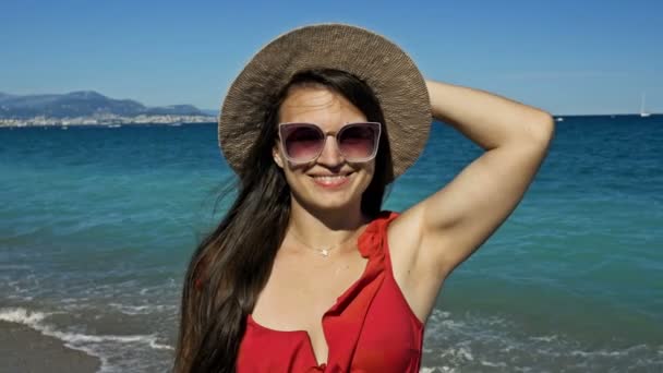 Vrolijke jonge vrouw met een zonnebril en een hoed staat aan de kust. Rotsachtige kusten en witte jachten zijn zichtbaar in de verte. — Stockvideo