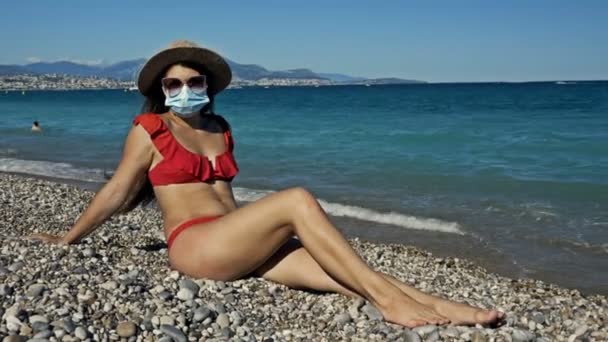 Νεαρή γυναίκα με ιατρική μάσκα και φαρδύ καπέλο κάνει ηλιοθεραπεία στην παραλία. Τουρισμός κατά τη διάρκεια του coronavirus. — Αρχείο Βίντεο