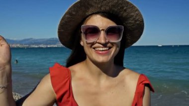 Güneş gözlüklü ve plaj şapkalı güzel genç bir kadın ellerini nazikçe sallıyor. O, deniz kıyısında duruyor. Arka planda kayalık bir sahil var, beyaz yatlar..