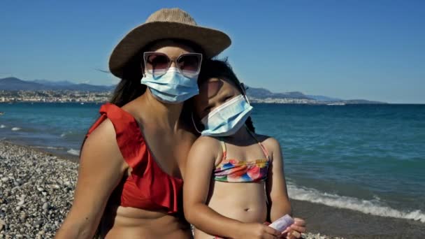 Молода жінка з маленькою донькою відпочиває на морському узбережжі. Обидва мають захисні маски. Туризм під час коронавірусу . — стокове відео