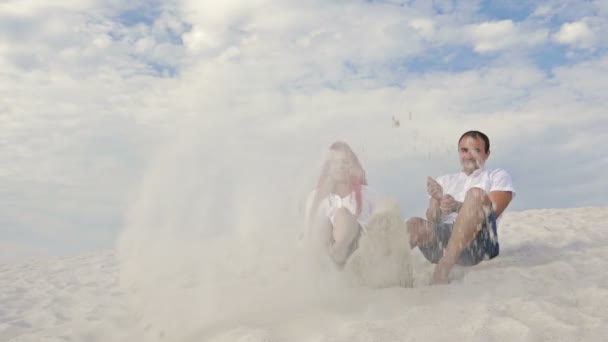Jovem casal alegre senta-se na areia branca e chuta a areia com os pés. Muito bom humor. Contra o fundo de um céu azul com nuvens brancas . — Vídeo de Stock
