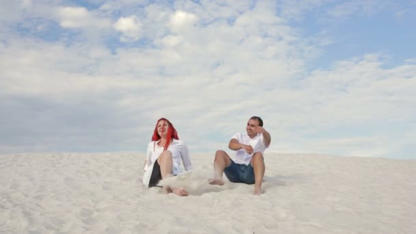 Joven pareja alegre se sienta en la arena blanca y patea la arena con sus pies. Buen humor. Sobre el fondo de un cielo azul con nubes blancas. — Vídeos de Stock